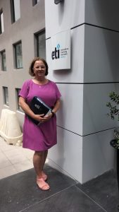 Na zdjęciu widzimy kobietę stojącą przed budynkiem, przy tablicy z napisem "eti executive training institute malta". Kobieta ubrana jest w fioletową sukienkę i różowe sandały, trzyma teczkę z logo "eti". Budynek, przy którym stoi, ma jasne ściany i kilka okien. Na zdjęciu widać również fragment doniczki z rośliną po prawej stronie.