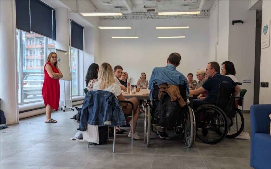 Grupa osób siedzi przy stole w jasnym, nowoczesnym pomieszczeniu podczas spotkania lub warsztatu. Część uczestników porusza się na wózkach inwalidzkich. Kobieta w czerwonej sukience stoi z boku z założonymi rękami i uśmiecha się. W tle duże okna z roletami.