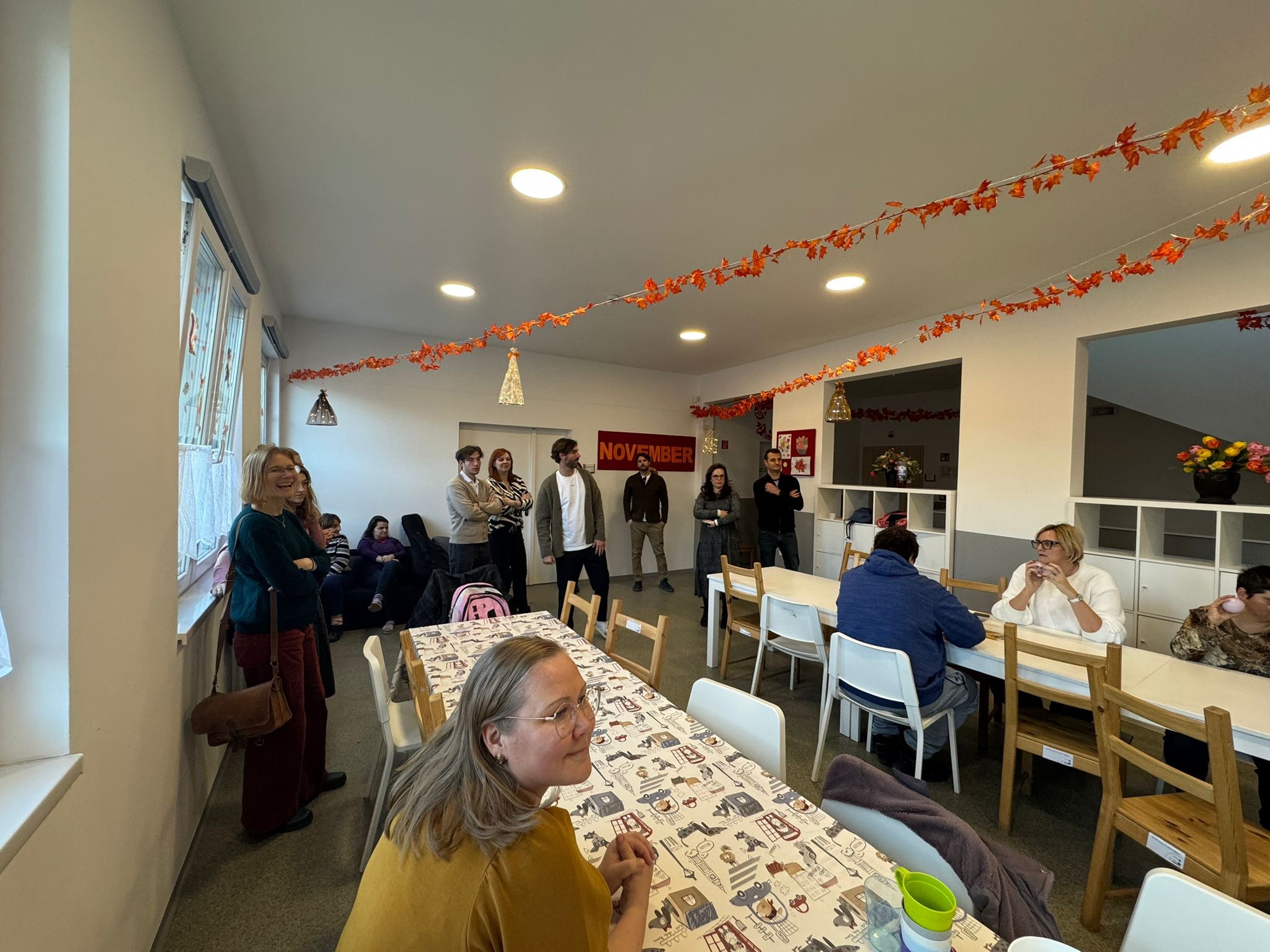 Zdjęcie wykonane wewnątrz jasnego, przestronnego pomieszczenia o charakterze świetlicy. Przez całą długość sali, pod sufitem, rozwieszone są girlandy z dekoracyjnych, pomarańczowych liści klonu, nawiązujące do jesieni. Na bocznej ścianie znajduje się duży czerwony transparent z napisem „NOVEMBER”. W pokoju ustawione są długie, białe stoły. Przy jednym z nich, na pierwszym planie, siedzi bokiem kobieta w żółtej bluzce i okularach. W głębi sali, przy kolejnych stołach, siedzą trzy osoby zajęte rozmową lub posiłkiem. Grupa osób znana z poprzednich zdjęć stoi wzdłuż ścian i pod oknami, przyglądając się wnętrzu i uczestnicząc w spotkaniu. Pomieszczenie jest dobrze doświetlone światłem dziennym wpadającym przez duże okna po lewej stronie oraz przez okrągłe plafony na suficie. Wnętrze jest czyste i uporządkowane, wyposażone w białe regały z przegrodami.