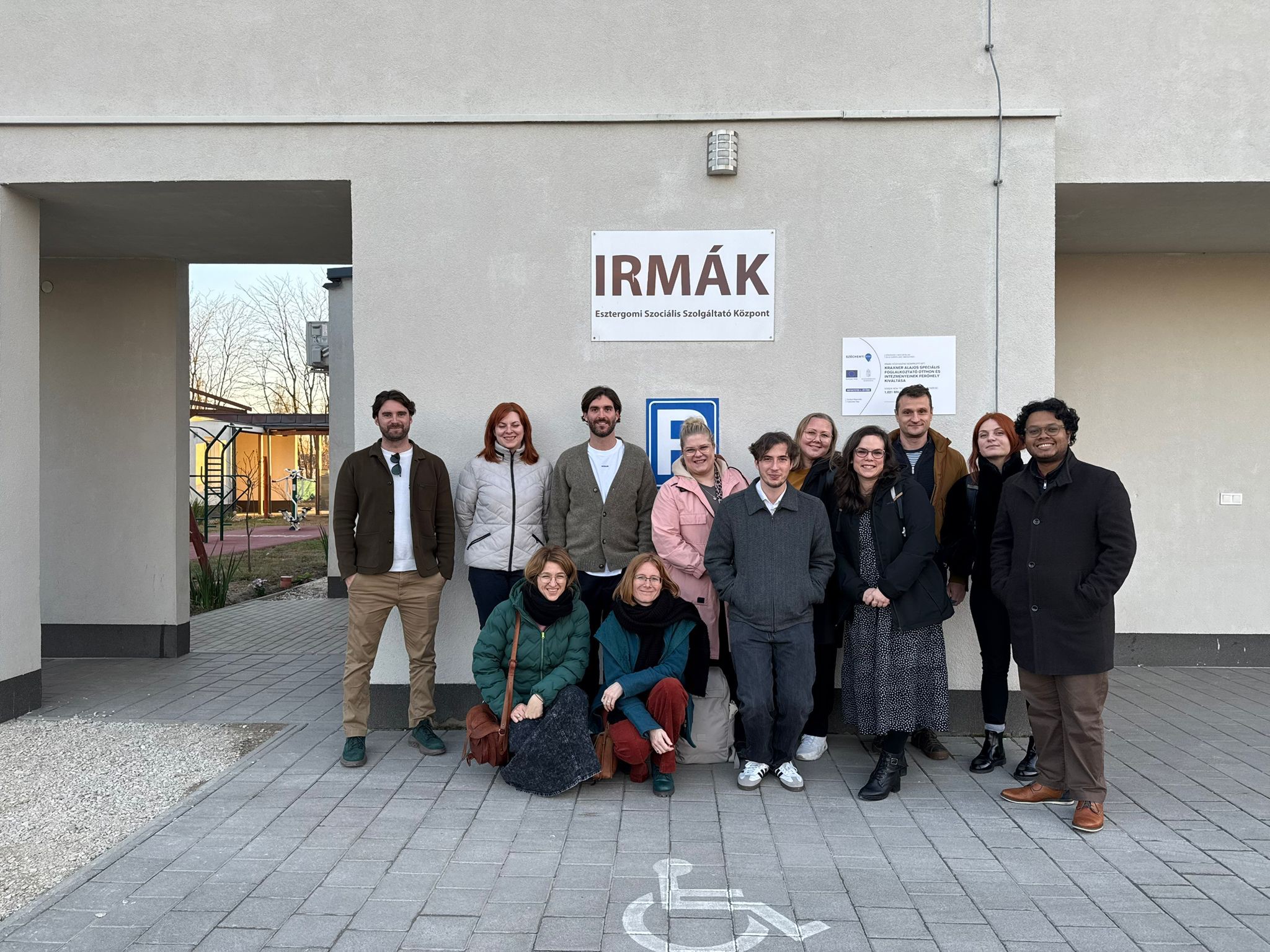 Grupa dwunastu osób stoi na zewnątrz przed jasną, gładką ścianą budynku. W centralnym punkcie, nad głowami zebranych, umieszczona jest biała tablica z brązowym napisem „IRMÁK Esztergomi Szociális Szolgáltató Központ”. Bezpośrednio pod tablicą, na elewacji, znajduje się niebieski znak informujący o miejscu parkingowym dla osób z niepełnosprawnościami. Grupa składa się z kobiet i mężczyzn w różnym wieku, ubranych w codzienne, jesienne stroje: kurtki, płaszcze i swetry. Dwie kobiety kucają w pierwszym rzędzie, pozostali uczestnicy stoją za nimi w luźnej formacji. Wszyscy patrzą w stronę aparatu i uśmiechają się. Podłoże stanowi równo ułożona szara kostka brukowa, na której w dolnej części kadru namalowany jest biały symbol wózka inwalidzkiego. Po lewej stronie, w głębi, znajduje się przejście prowadzące na teren z ogrodem i urządzeniami rekreacyjnymi.