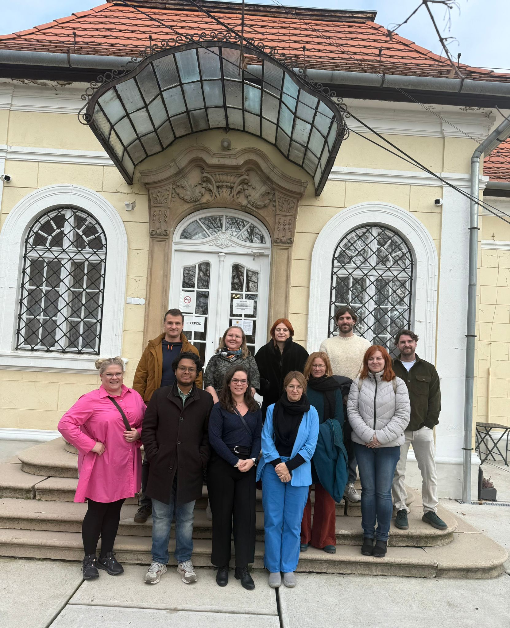Fotografia przedstawia tę samą grupę osób, tym razem pozującą na szerokich, kamiennych schodach przed wejściem do klasycznego, żółtego budynku. Budynek charakteryzuje się dużymi, białymi, łukowatymi oknami z ozdobnymi szprosami. Nad białymi, dwuskrzydłowymi drzwiami wejściowymi znajduje się dekoracyjny, półokrągły daszek wykonany ze szkła i ciemnego metalu. Uczestnicy spotkania są ustawieni w trzech rzędach. Po lewej stronie, na dole schodów, wyróżnia się kobieta w jaskraworóżowej koszuli. Inna osoba, stojąca w środku pierwszego rzędu, ma na sobie niebieski garnitur i czarny szal. Pozostali członkowie grupy noszą ubrania w kolorach ziemi, czerni i dżinsu. Cała grupa promienieje radością. Nad budynkiem znajduje się dach z czerwonej dachówki, a po prawej stronie biegnie srebrna rura spustowa.